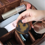 Hand holding a tortoiseshell compact mirror over a makeup bag filled with cosmetic items.
