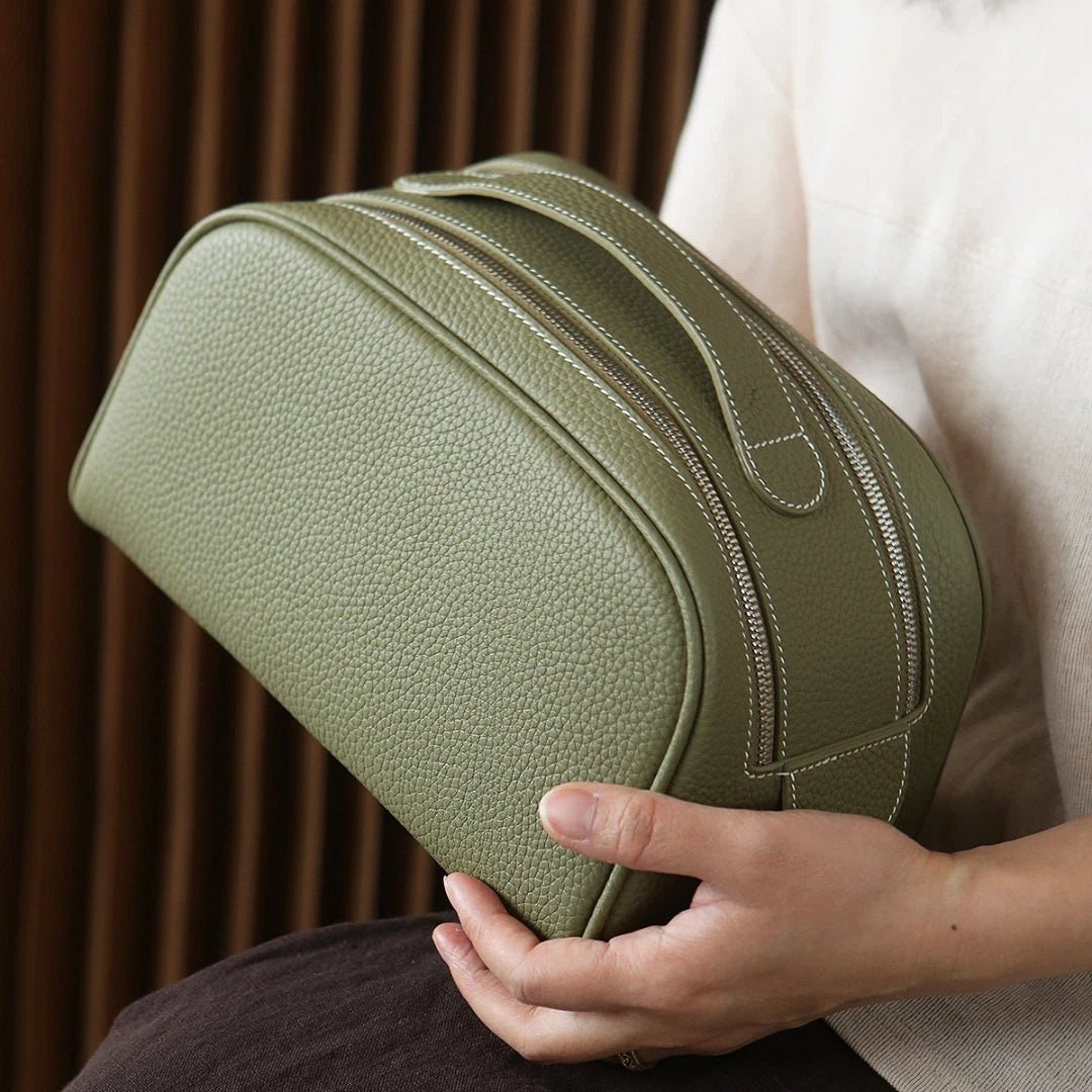 Person holding a green leather toiletry bag against a neutral background