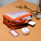 An orange cosmetic bag with a translucent frosted top panel, shown open with makeup items partially pulled out, placed on a tan surface next to a glass of water.