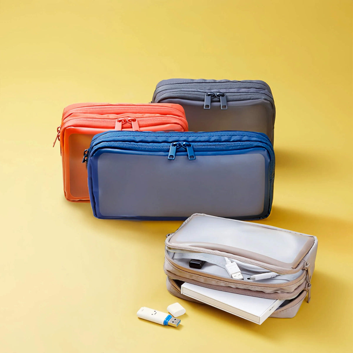 Four zippered travel bags in different colors are arranged on a yellow studio background. The front-most bag is beige and shown open with a notebook, charging cable, and small items inside.