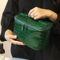 A person in a black top holds a glossy green cosmetic bag with silver zippers. The person's hands and part of their torso are visible, with a small bottle at the bottom left.
