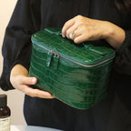 A person in a black top holds a glossy green cosmetic bag with silver zippers. The person's hands and part of their torso are visible, with a small bottle at the bottom left.