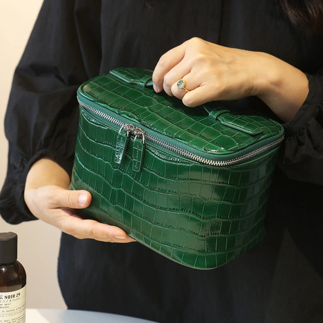 A person in a black top holds a glossy green cosmetic bag with silver zippers. The person's hands and part of their torso are visible, with a small bottle at the bottom left.