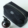 A black nylon cosmetic pouch with silver zippers and a small oval 'amplitude' logo patch, shown on a white background next to a brown pump bottle.