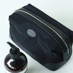 A black nylon cosmetic pouch with silver zippers and a small oval 'amplitude' logo patch, shown on a white background next to a brown pump bottle.