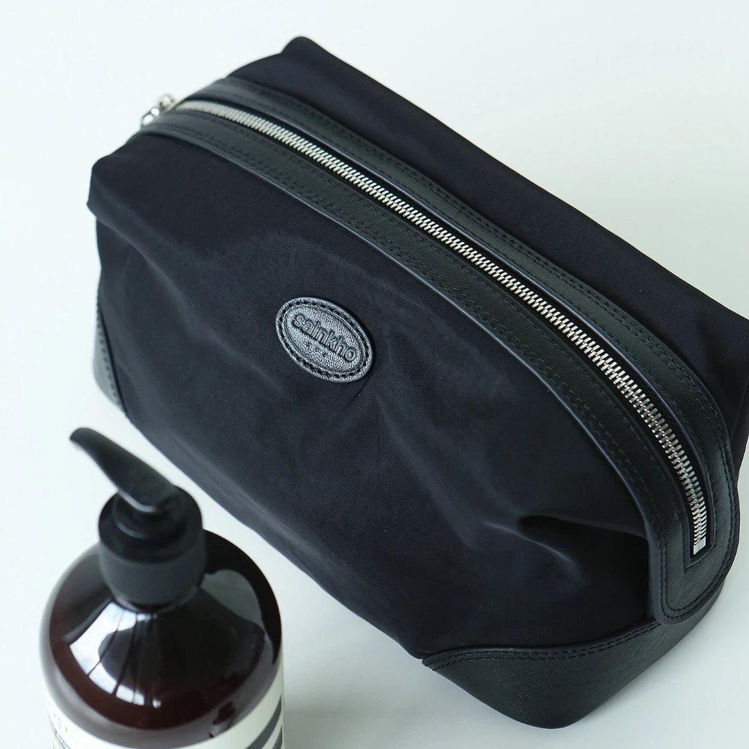 A black nylon cosmetic pouch with silver zippers and a small oval 'amplitude' logo patch, shown on a white background next to a brown pump bottle.
