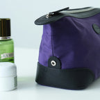 A purple nylon cosmetic bag with black leather trim, shown in a close-up product shot on a white surface with a La Mer bottle and a white jar as styled props in the background.