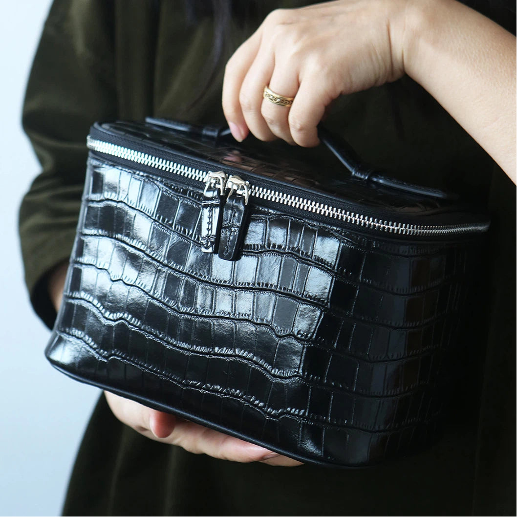 A person holding a glossy black makeup bag with a top handle and silver zipper closure.