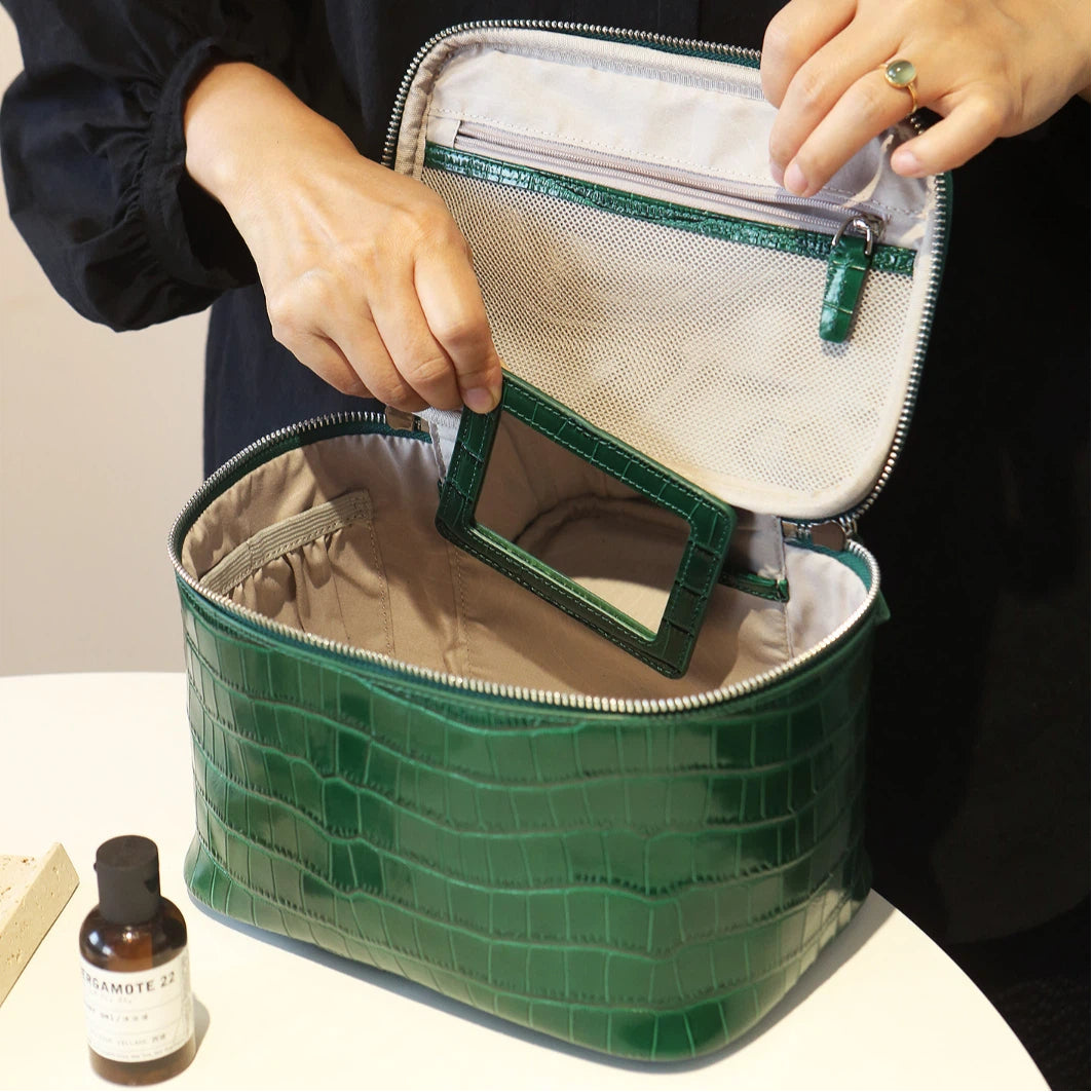 A green cosmetic bag is shown open with a person's hands inside, revealing a light gray interior, zippered compartments, and a small mirror. A small bottle sits on the table beside it.