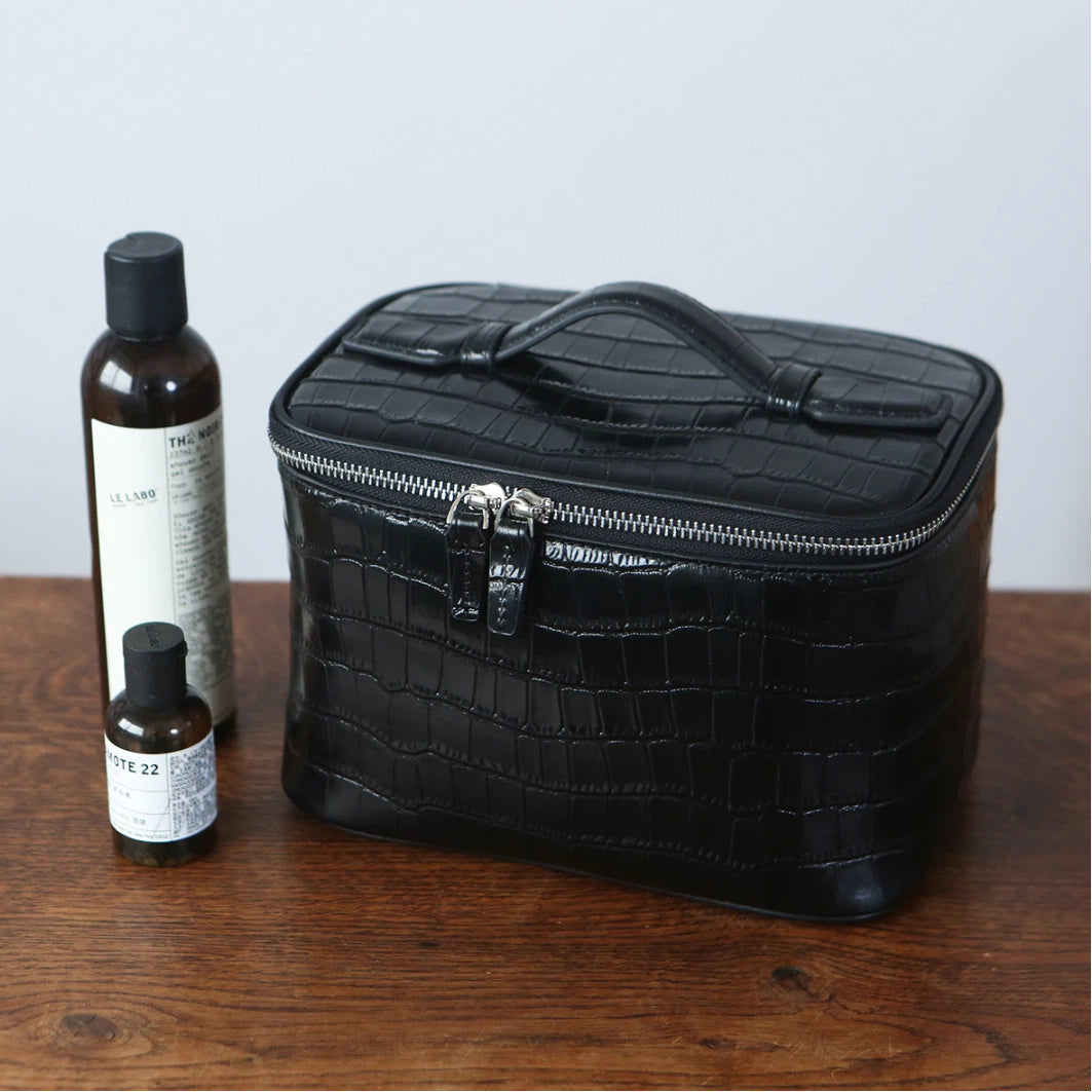 A black zippered cosmetic bag with a top handle sits on a wooden table next to two dark bottles with white labels.