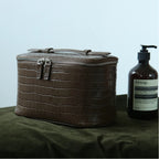 A brown zippered cosmetic bag sits on a dark olive green cloth next to an Aesop pump bottle against a light blue-gray background.