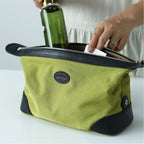 A green canvas toiletry bag with black trim, shown with a hand placing a white item inside and holding a green bottle. The bag has a circular black logo patch on the front.