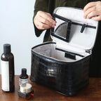 A black cosmetic bag is shown open on a wooden table, with a person's hands holding the inner lid. The bag has a light gray interior and a clear zippered pocket on the inside lid.