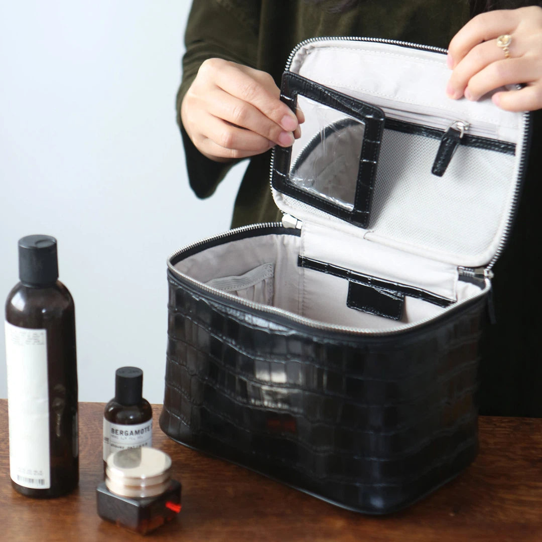 A black cosmetic bag is shown open on a wooden table, with a person's hands holding the inner lid. The bag has a light gray interior and a clear zippered pocket on the inside lid.