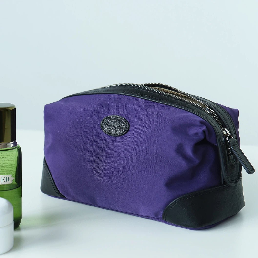 A purple cosmetic pouch with black leather trim and a top zipper, shown on a light gray background with a green bottle partially visible on the left.