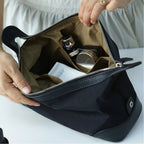 A black nylon cosmetic bag with a brown interior organizer, shown being opened by hands to reveal toiletry items inside.