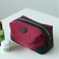 A burgundy nylon cosmetic pouch with black trim and a silver zipper, featuring a circular black logo patch on the front, displayed on a white surface next to skincare items.