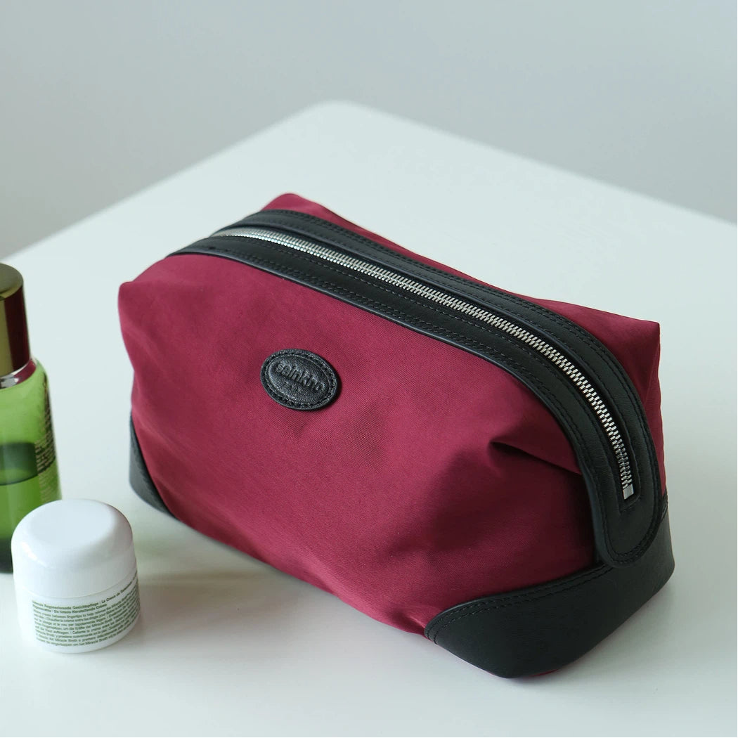 A burgundy nylon cosmetic pouch with black trim and a silver zipper, featuring a circular black logo patch on the front, displayed on a white surface next to skincare items.