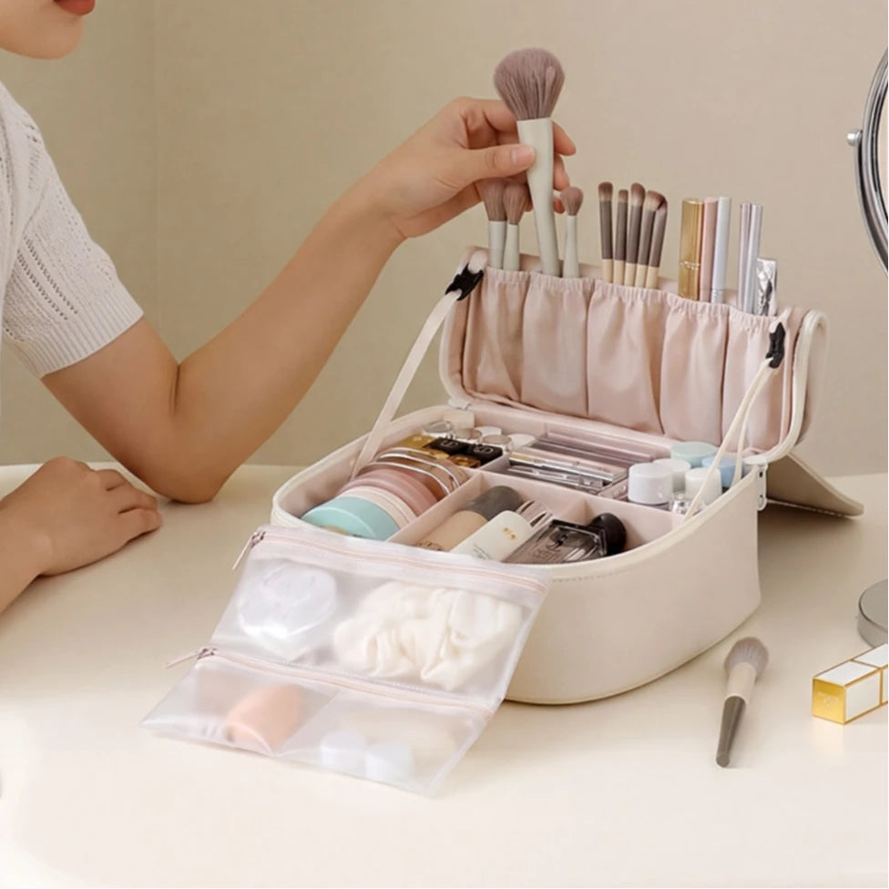 A beige makeup organizer case on a light table, shown open with brushes, cosmetics, and a clear zip pouch in front. A person's hand is holding a makeup brush above the organizer, and part of a mirror is visible on the right.