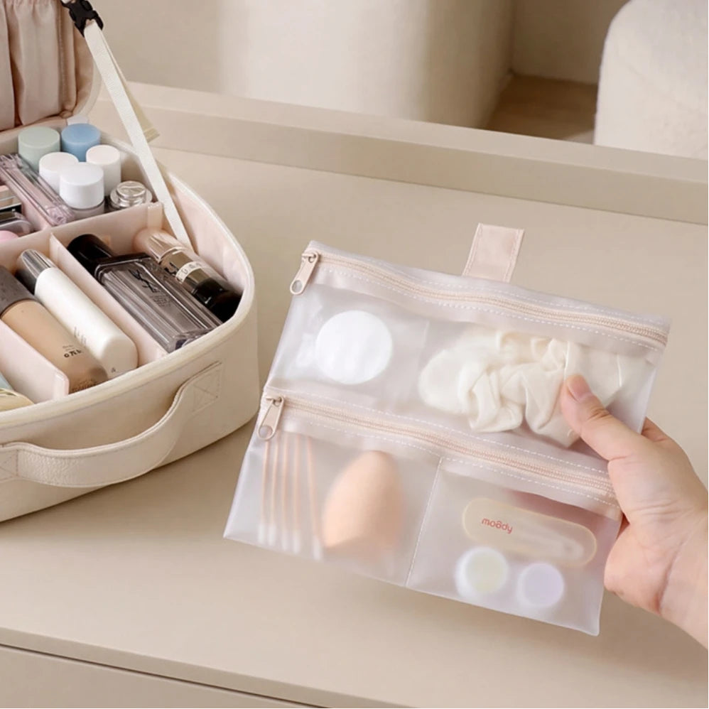 A hand holds a clear zippered cosmetic organizer pouch with multiple compartments, shown beside an open beige makeup bag on a beige tabletop.