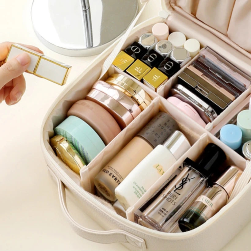 A beige makeup organizer case with multiple compartments filled with cosmetics, shown open on a light background. A hand is holding a small gold-framed item near the case.