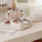 A cream makeup organizer case is shown open on a vanity table, filled with cosmetics and brushes. A second closed matching case sits nearby, and part of a person's arm is visible holding the open case.