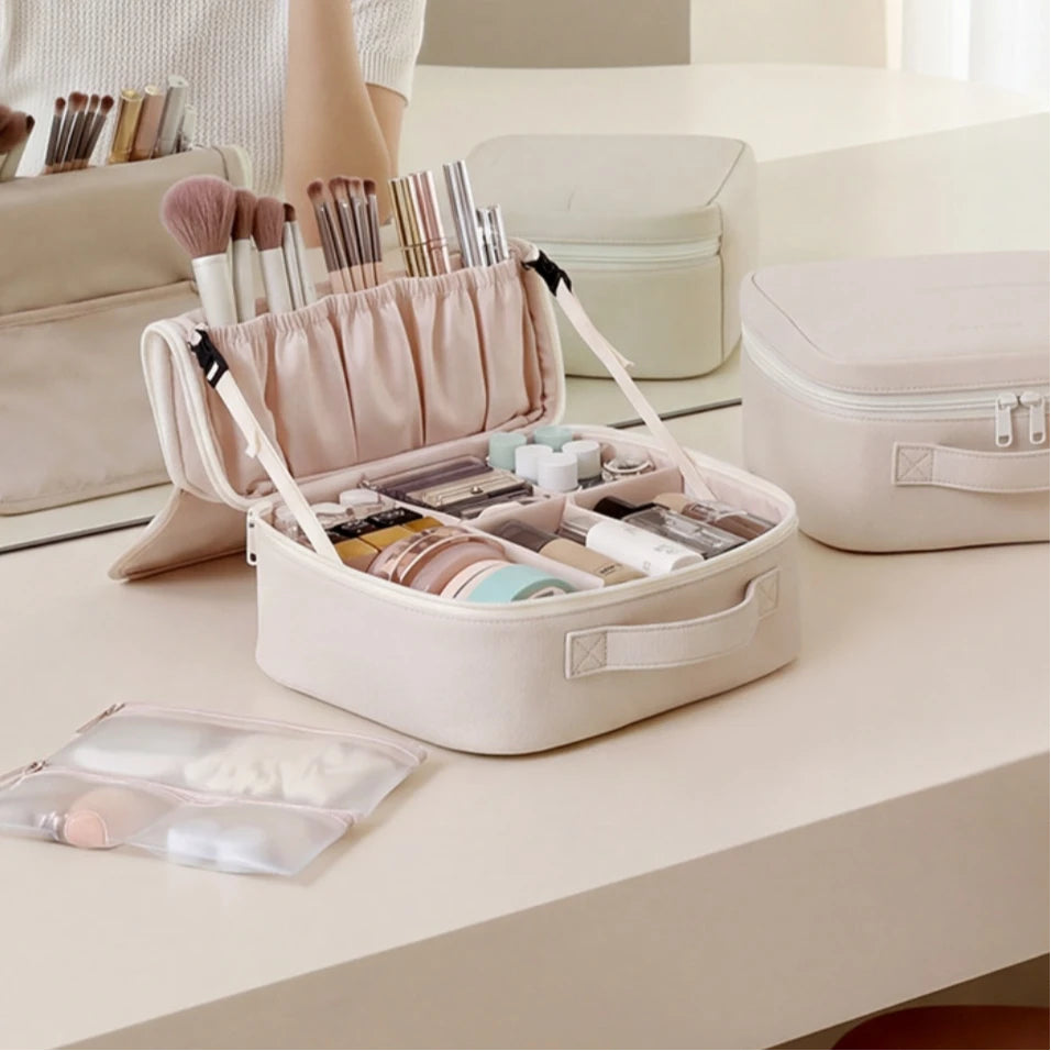 A cream makeup organizer case is shown open on a vanity table, filled with cosmetics and brushes. A second closed matching case sits nearby, and part of a person's arm is visible holding the open case.